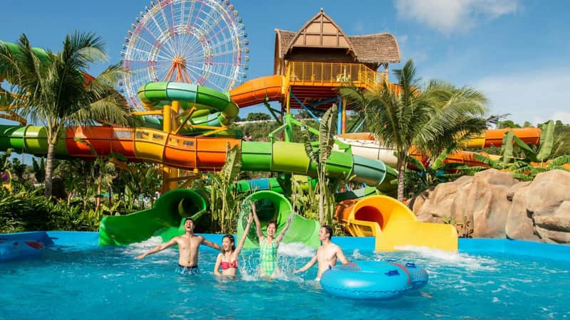 Four people enjoying a pool at a waterpark with colorful slides, tropical plants, and a Ferris wheel in the background.
