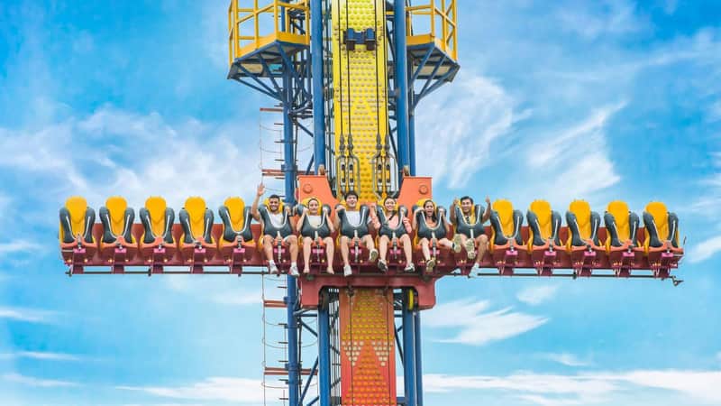A group of people seated on a vertical drop amusement ride, smiling and raising their hands under a bright blue sky.