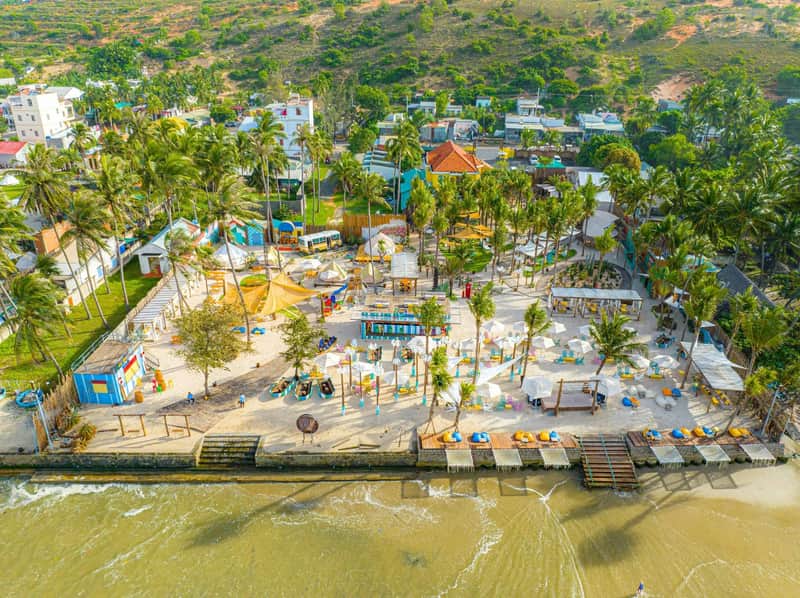 A vibrant tropical beach area with palm trees, colorful umbrellas, buildings, and people enjoying the seaside setting.