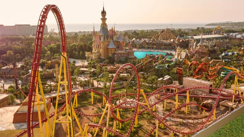A red and yellow rollercoaster in the foreground with a castle and theme park in the background under a sunny sky.