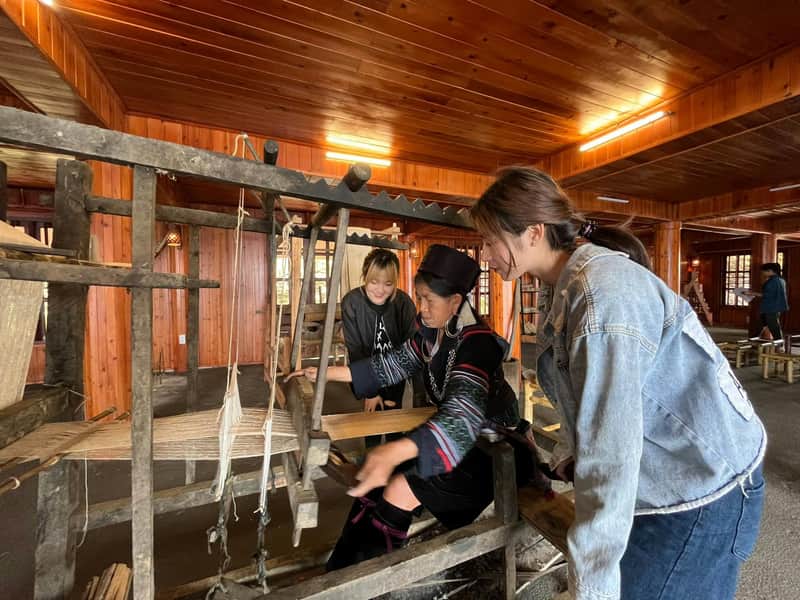 Three women in a wooden interior operate a traditional loom, with one demonstrating and the others observing attentively.