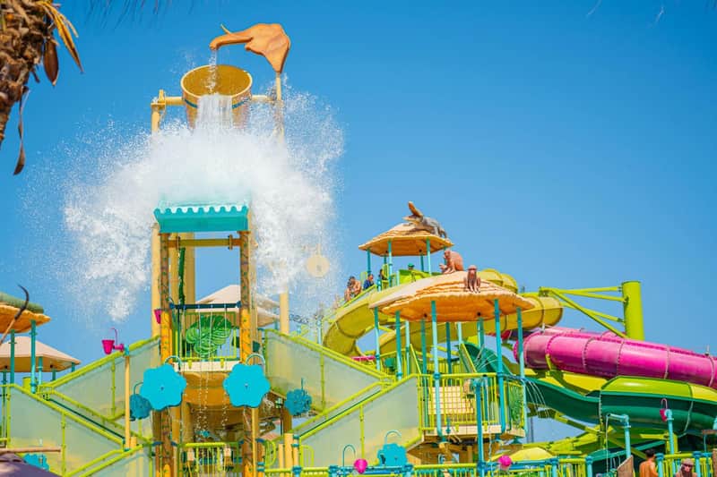 A colorful water playground with slides, a large tipping bucket, and playful decorations under a clear blue sky.