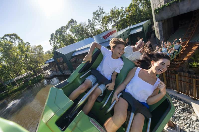 People enjoying a thrilling rollercoaster ride with a riverside setting and forested background on a sunny day.