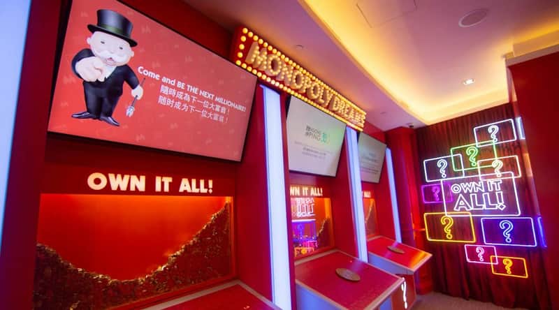 A colorful Monopoly-themed interactive exhibit with neon lights, screens, and a sign reading "Own It All!" in bold letters.