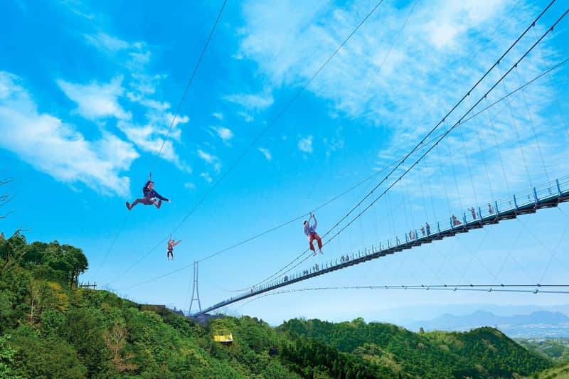 People zip-lining over a lush green forest near a suspension bridge under a bright blue sky with scattered clouds.