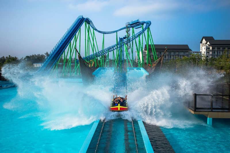 A log flume ride splashing into water with a vibrant roller coaster in the background under a clear blue sky.