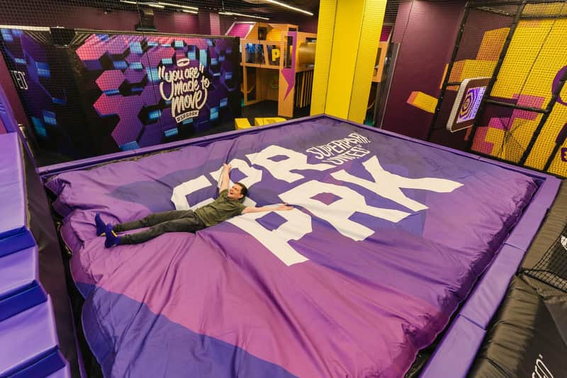 A person lies on a purple crash mat with white text in an indoor trampoline park with colorful walls and cushioned surfaces.