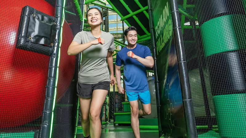 Two people running and smiling inside an indoor obstacle course with green and black structures.