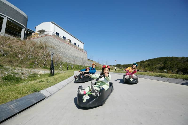 Children riding downhill on luge carts on a paved track under a clear blue sky, with a building and greenery in the background.