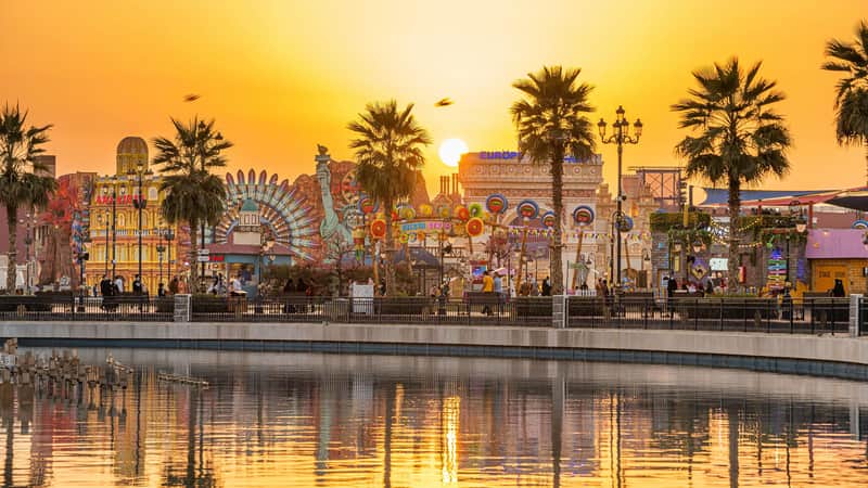 Amusement park at sunset with colorful structures, palm trees, and people walking along a waterfront. Reflections in the water.