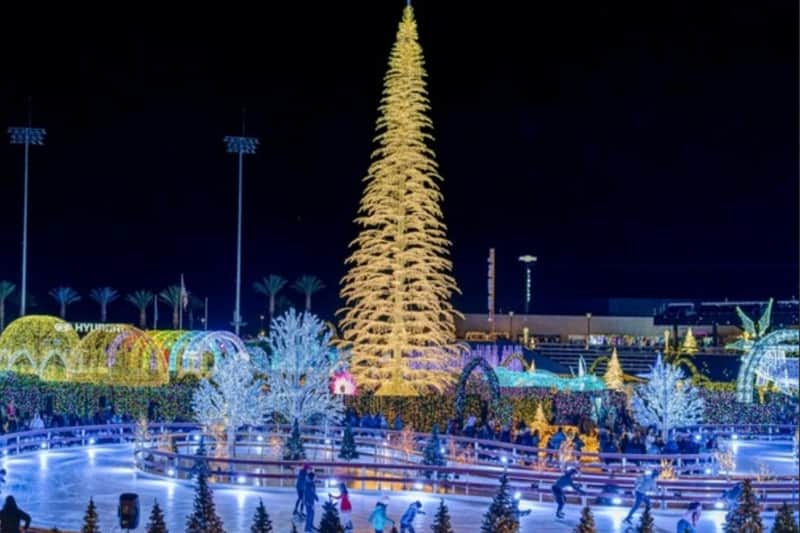 A brightly lit outdoor ice skating rink surrounded by festive holiday lights and decorations, including a large illuminated tree.