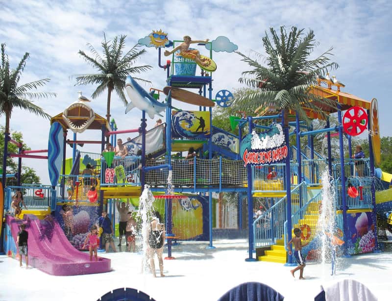 Children playing at a colorful water park featuring slides, climbing structures, fountains, and palm trees under a partly cloudy sky.