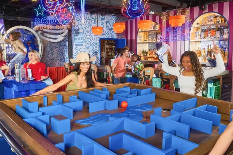A lively bar scene with people playing a large tabletop maze game and others enjoying drinks under colorful neon lights.