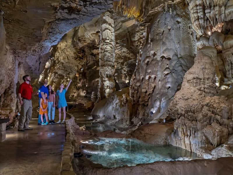 Natural Bridge Caverns Park Tickets (San Antonio) Five people exploring a cave with stalactites, stalagmites, and a shallow pond. One person points toward the cave formations.