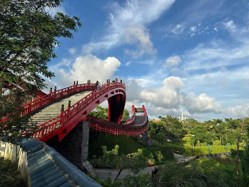 A red arched pedestrian bridge with railings spans a green park under a blue sky with clouds.