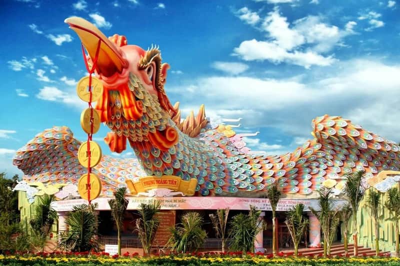 A large, colorful dragon sculpture with coins hanging from its mouth, vibrant scales, and foliage in the foreground under a blue sky.