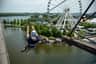 Person ziplining over a body of water with a Ferris wheel and green trees in the background.