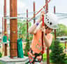 A child wearing a white helmet and harness navigates a ropes course outdoors, holding onto a red safety line.