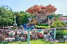 Amusement park with colorful rides, large tree structure, and people enjoying various attractions under a clear blue sky.