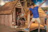 A child in a blue shirt and beige shorts jumps in front of a wooden playhouse structure with other children nearby.