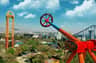 Amusement park ride in motion, spinning high above a cityscape with buildings, trees, and a blue sky with clouds in the background.