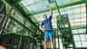 Une personne en tenue de sport tendant les bras vers le haut dans une aire de jeux couverte comportant des éléments structurels verts et un plafond en verre.