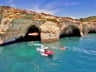 A scenic coastal view featuring rocky cliffs with sea caves and several boats on turquoise water under a clear blue sky.