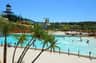 A water park with people swimming in a wave pool, surrounded by palm trees and water slides under a clear blue sky.