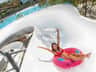Woman in a red bikini on a pink inflatable tube sliding down a white water slide, with a pool and trees in the background.