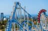 A blue and white rollercoaster with loops and drops, with a cityscape and ocean in the background under a clear sky.