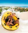 Three people on a yellow inflatable raft ride down a water slide with splashing water, cityscape and ocean in the background.