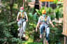 Two people wearing helmets and safety gear ride bicycles suspended on a zipline in a lush, forested setting.