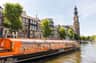 Orange canal cruise boat docked in an Amsterdam canal, with historic buildings and a church tower in the background.