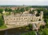 Aerial view of a large stone castle with multiple towers, surrounded by greenery, water, and a distant townscape.