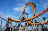 A roller coaster with an orange and yellow loop, carrying people, set against a blue sky with palm trees in the background.