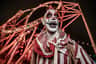 A person in clown makeup and costume standing in front of an illuminated roller coaster at night.