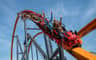 People on a rollercoaster with a dragon-themed front, raising their arms and smiling under a clear blue sky.