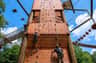 Two people climbing a tall wooden climbing wall outdoors, with bright sky and ropes elements visible in the background.