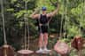 A person wearing a safety harness is balancing on suspended wooden platforms in an outdoor adventure park with trees in the background.