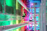 A person in a harness traverses a high indoor ropes course illuminated by colorful lights in a large industrial-style space.
