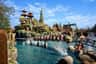 A themed rollercoaster passes over water in a fantasy village setting with rocky structures under a clear blue sky.