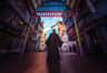 A person in a long black robe stands facing a dragon sculpture that is breathing fire in a themed alleyway.
