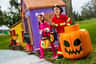 Two children in Halloween costumes, a pink cat and a firefighter, holding candy buckets in front of a playful, spooky house.