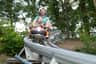 A man and child ride together on a roller coaster through a wooded area, both appearing excited and smiling.