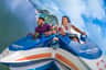 People smiling and enjoying a rollercoaster ride, with water and trees in the background.