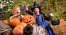 A child smiles surrounded by pumpkins and a LEGO jack-o’-lantern, with festive flags and trees in the background.