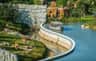 A boat with passengers travels along a canal next to a zoo enclosure with tigers and rocks in a landscaped area.