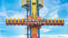 People seated on a vertical amusement ride, ascending against a blue sky, with visible support structure and safety harnesses.