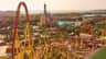 A theme park with a red and yellow roller coaster, a castle-like structure, and other attractions under a sunny sky.