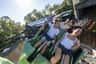 People enjoying a thrilling rollercoaster ride with a riverside setting and forested background on a sunny day.